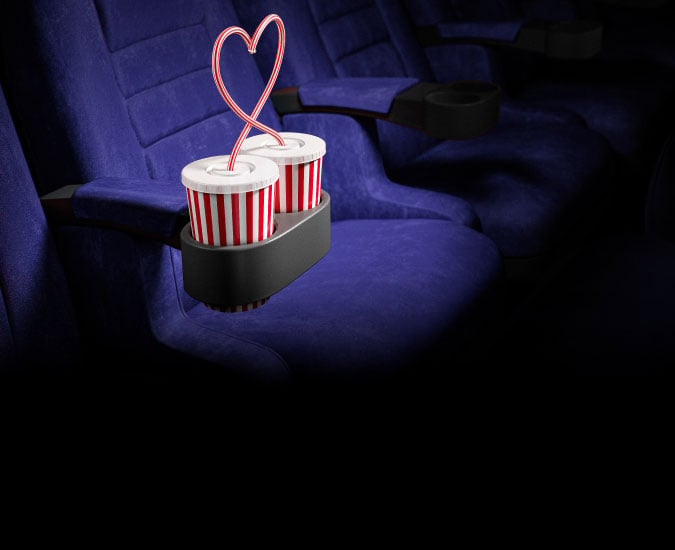 Two drinks beside each other in cup holders of movie thetare chairs.  Their straws are creating a heart shape.