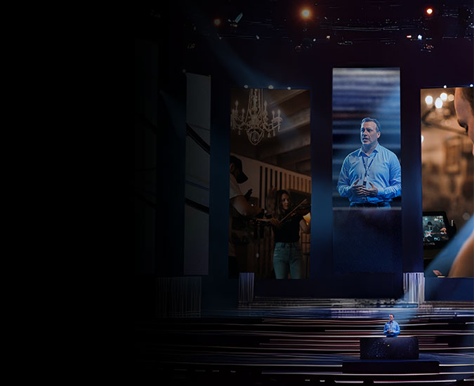 A person presenting on stage with large vertical screens behind him projecting images of him and the subject matter.