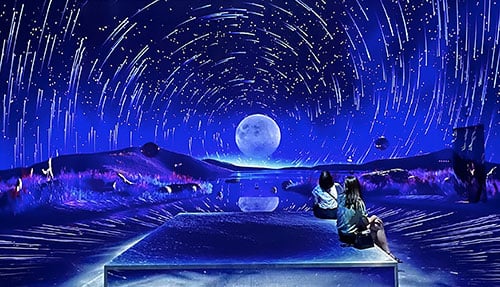 2 people sitting in an immersive room with swirling stars and a full moon against a deep blue sky projected around them.