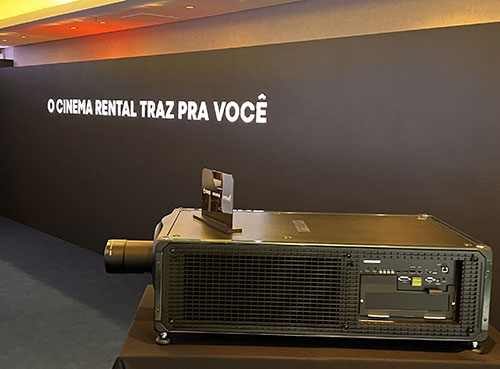 The side view of a Cinema projector in front of a wall with a sign that says "O CINEMA RENTAL TRAZ PRA VOCE