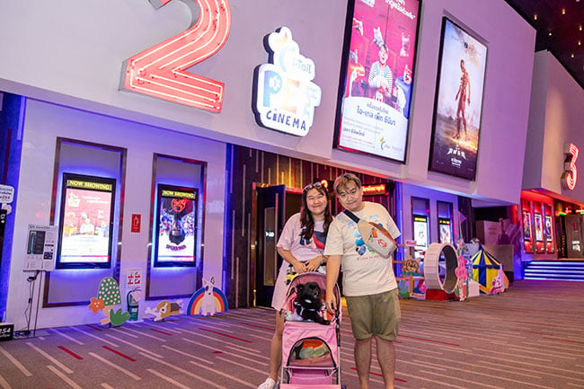A man and a woman stand with their black dog that is in a pink stroller outside of an i-Tail Pet Cinema.