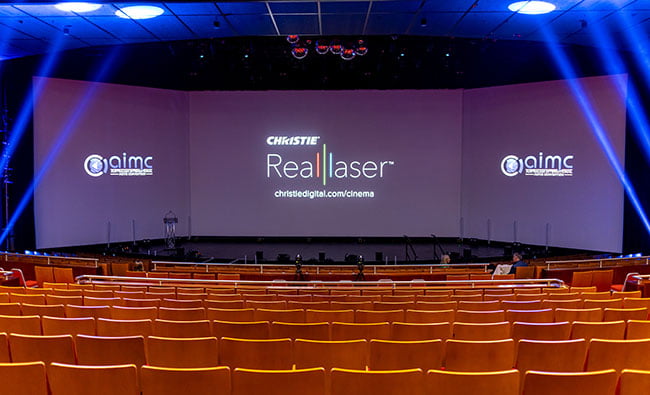 Three screens are projected with text on a large auditorium stage.