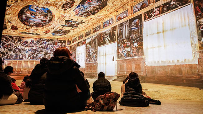 People admire projections of a large painting in an art museum.