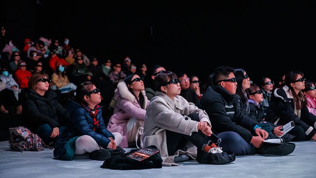 People wearing 3D glasses are seated on the floor watching a performance.