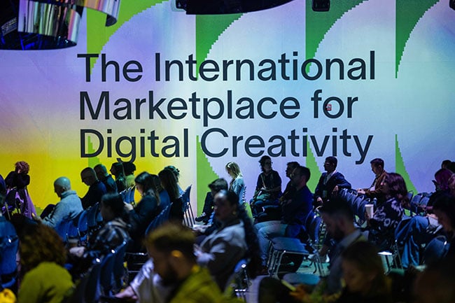 People sitting in chairs in a large room with the text “The International Marketplace for Digital Creativity” projected onto the wall.