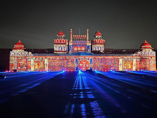 The walls of a gigantic monument is colorfully lit by projectors at night.