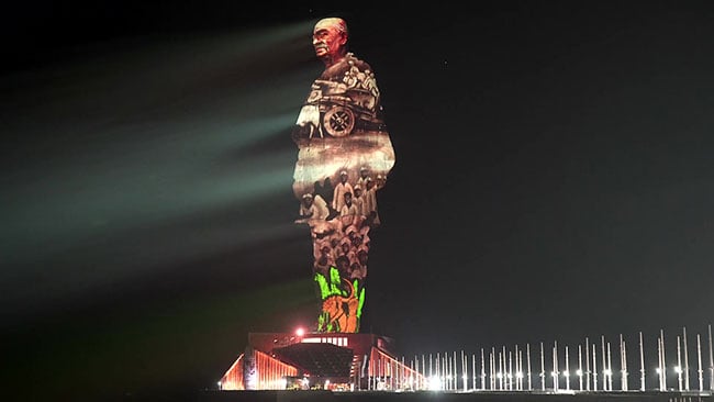 A large statue lit by multiple projectors at night.