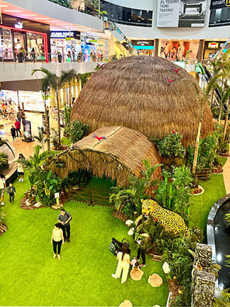 A thatched geodesic dome is surrounded by plants and artificial turf inside a shopping mall. 