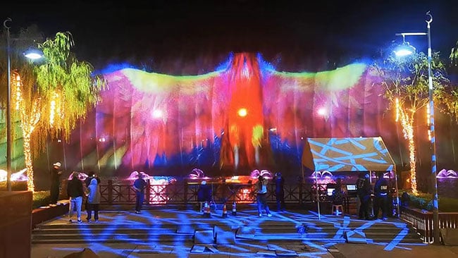 An image of a colorful bird is projected onto a large water screen.