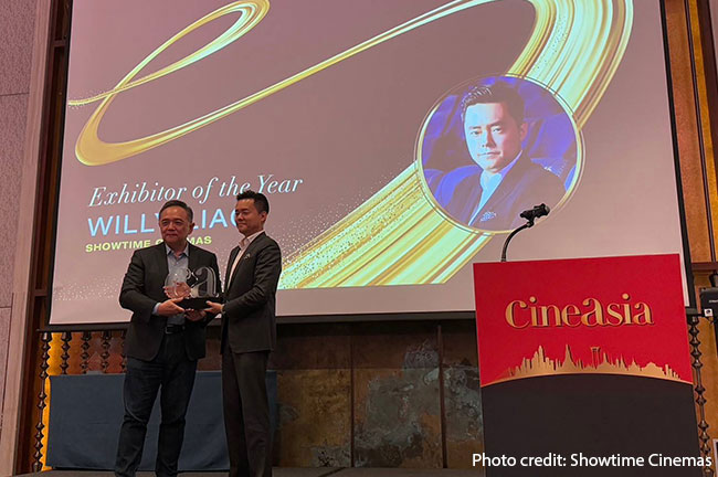 Two men stand on a large stage holding an award.