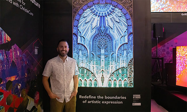 A man stands in front of a piece of art that is projection mapped in bright colors.