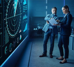 Two people standing in front of a large screen in a control room, gesturing to a clipboard one of them is holding and having a discussion.